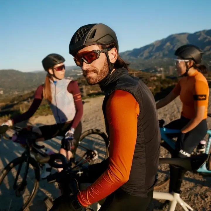 Homem de roupas de ciclista visto na frente de duas mulheres com roupas de ciclistas também; todos estão montados em bicicletas e usando óculos especiais; percebe-se uma paisagem montanhosa e um céu azul limpo no fundo. Os capacetes de todos são impressos em 3D.