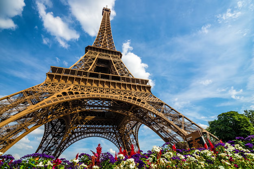 Essa imagem mostra de forma ilustrativa uma foto com vista térrea da Torre Eiffel, onde é dia e pode-se ver um céu azul com poucas nuvens brancas e flores nos pés da torre.