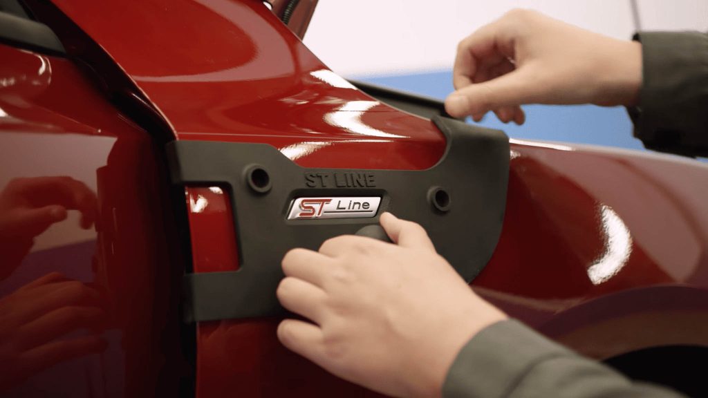 Duas mãos encaixando uma peça impressa preta em um automóvel vermelho; não podemos ver o automóvel todo, apenas uma parte pequena ampliada.