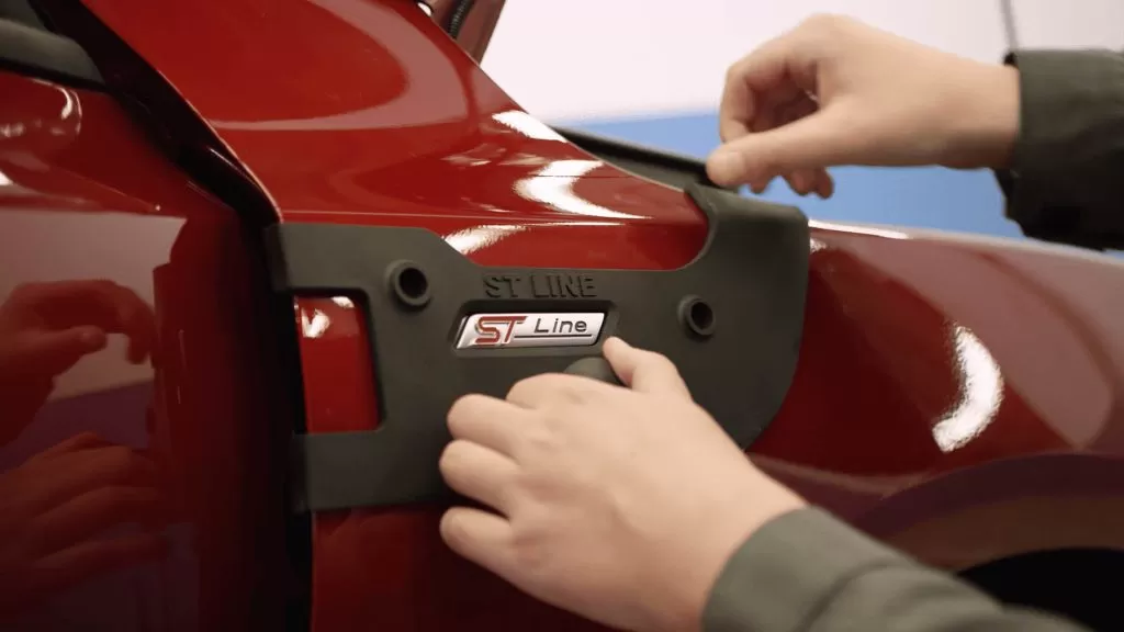 Duas mãos encaixando uma peça impressa preta em um automóvel vermelho; não podemos ver o automóvel todo, apenas uma parte pequena ampliada.